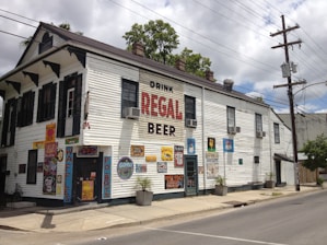 A two-story white building with several advertisements and colorful signs on the walls. It has a large sign that reads 'Drink Regal Beer.' The structure has multiple air conditioners, dark shutters, and posters displaying menu items like waffles and po'boys. There are plants in pots along the sidewalk and a street with power lines overhead.