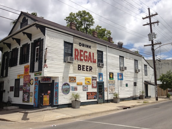A two-story white building with several advertisements and colorful signs on the walls. It has a large sign that reads 'Drink Regal Beer.' The structure has multiple air conditioners, dark shutters, and posters displaying menu items like waffles and po'boys. There are plants in pots along the sidewalk and a street with power lines overhead.
