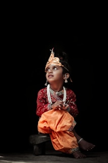 A child dressed in traditional attire with a decorated headpiece and jewelry, sitting on a small wooden stool against a dark background. The child gazes upwards with an expression of curiosity and wonder, wearing a red embroidered top and orange pants. Pearl necklaces and anklets enhance the vibrant cultural costume.