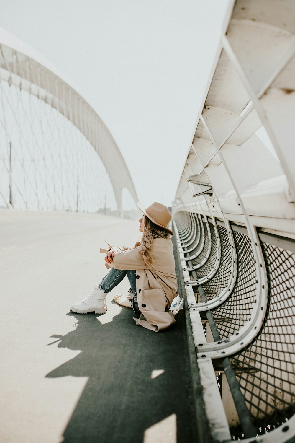 woman in brown leather jacket and blue denim jeans sitting on white metal fence during daytime