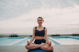 A person is seated in a yoga pose on a mat next to an outdoor swimming pool. The background features a cloudy sky and a glass barrier, with plants visible along the poolside.