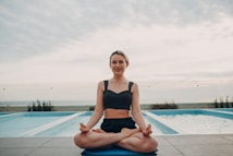 A person is seated in a yoga pose on a mat next to an outdoor swimming pool. The background features a cloudy sky and a glass barrier, with plants visible along the poolside.