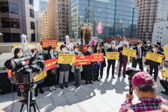 A group of people are gathered outdoors in an urban setting, holding signs that advocate against racism, sexism, and violence. They are wearing masks and standing in front of a statue. A video camera is focused on the group, and another person appears to be capturing the scene with a handheld device. Skyscrapers form the backdrop, and a pink flag with a symbol is visible.