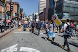 A group of people are gathered on a city street, participating in a protest or public demonstration. Some hold signs with messages, and an individual in a bright orange vest stands out among the crowd. Tall buildings and traffic signals are visible in the background, alongside urban elements like street signs and a crosswalk. The crowd appears diverse and engaged, suggesting a communal gathering.