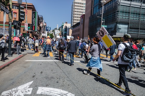 A group of people are gathered on a city street, participating in a protest or public demonstration. Some hold signs with messages, and an individual in a bright orange vest stands out among the crowd. Tall buildings and traffic signals are visible in the background, alongside urban elements like street signs and a crosswalk. The crowd appears diverse and engaged, suggesting a communal gathering.