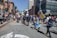 A group of people are gathered on a city street, participating in a protest or public demonstration. Some hold signs with messages, and an individual in a bright orange vest stands out among the crowd. Tall buildings and traffic signals are visible in the background, alongside urban elements like street signs and a crosswalk. The crowd appears diverse and engaged, suggesting a communal gathering.