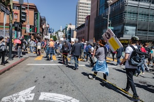 A group of people are gathered on a city street, participating in a protest or public demonstration. Some hold signs with messages, and an individual in a bright orange vest stands out among the crowd. Tall buildings and traffic signals are visible in the background, alongside urban elements like street signs and a crosswalk. The crowd appears diverse and engaged, suggesting a communal gathering.