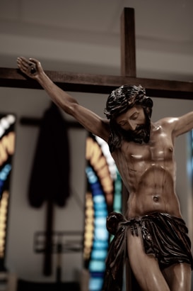 A detailed wooden sculpture of a man crucified, with arms outstretched on a cross. The expression on his face is somber, with closed eyes and a hint of suffering. In the background, there are colorful, blurred stained glass windows, adding a vibrant contrast to the muted tones of the sculpture.