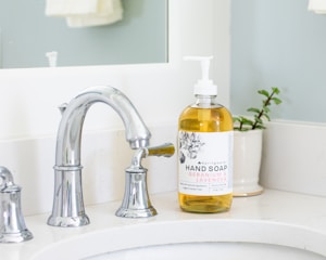 A modern bathroom sink featuring a sleek chrome faucet is complemented by a stylish bottle of geranium and lavender hand soap placed nearby. The label on the soap bottle is elegantly designed, and a small potted plant adds a touch of greenery next to the sink.