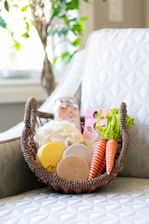 A wicker basket holds a variety of natural and handmade bath products, including bath bombs, salts, and soaps. Two realistic carrot-shaped soaps or decorations add a unique touch. The basket is placed on a patterned white cushioned chair, with a blurred leafy plant in the background enhancing the serene setting.