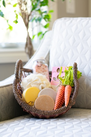 A wicker basket holds a variety of natural and handmade bath products, including bath bombs, salts, and soaps. Two realistic carrot-shaped soaps or decorations add a unique touch. The basket is placed on a patterned white cushioned chair, with a blurred leafy plant in the background enhancing the serene setting.