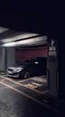 A darkly lit parking garage with a grey BMW car parked in a designated parking spot. The concrete pillar next to the car has signage in Turkish indicating 'Office Entrance'. The floor markings and lighting create a moody atmosphere.