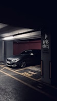 A darkly lit parking garage with a grey BMW car parked in a designated parking spot. The concrete pillar next to the car has signage in Turkish indicating 'Office Entrance'. The floor markings and lighting create a moody atmosphere.