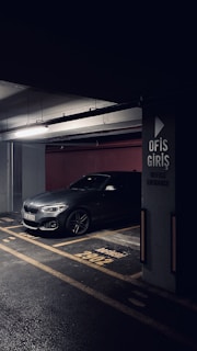 A darkly lit parking garage with a grey BMW car parked in a designated parking spot. The concrete pillar next to the car has signage in Turkish indicating 'Office Entrance'. The floor markings and lighting create a moody atmosphere.