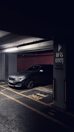 A darkly lit parking garage with a grey BMW car parked in a designated parking spot. The concrete pillar next to the car has signage in Turkish indicating 'Office Entrance'. The floor markings and lighting create a moody atmosphere.