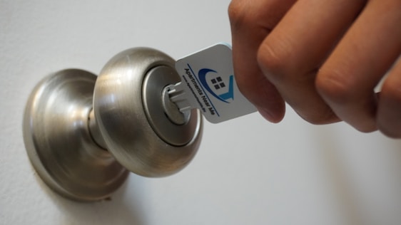 A hand is seen holding a key card, inserting it into a modern doorknob with a built-in lock. The key card has a logo and some text printed on it.