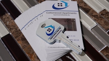 A stack of brochures for Cottonwood Apartments is placed on a tiled surface. On top of the brochures, there is a key with a logo that features a house and the words 'Apartments Near Me'. The brochures include contact information and a picture of the apartment interior.