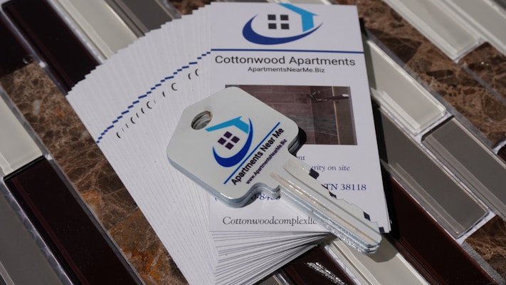 A stack of brochures for Cottonwood Apartments is placed on a tiled surface. On top of the brochures, there is a key with a logo that features a house and the words 'Apartments Near Me'. The brochures include contact information and a picture of the apartment interior.