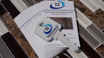 A stack of brochures for Cottonwood Apartments is placed on a tiled surface. On top of the brochures, there is a key with a logo that features a house and the words 'Apartments Near Me'. The brochures include contact information and a picture of the apartment interior.