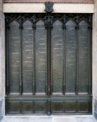 A large, ornate bronze door is adorned with detailed engravings and inscriptions. The door features several vertical panels framed by decorative columns and topped with an ornate frieze. Above the panels, an inscribed Latin text sits below a small sculptural element.