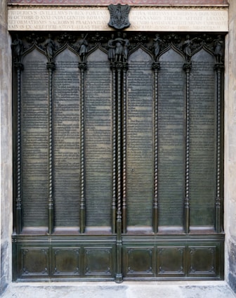A large, ornate bronze door is adorned with detailed engravings and inscriptions. The door features several vertical panels framed by decorative columns and topped with an ornate frieze. Above the panels, an inscribed Latin text sits below a small sculptural element.