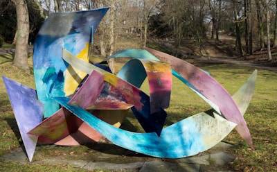 A colorful abstract metal sculpture is set outdoors in a park surrounded by trees. The sculpture consists of various curved and angular shapes painted in vibrant shades of blue, yellow, pink, and purple, with a dynamic and intricate design.