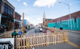 An outdoor urban area with a wooden picket fence enclosing a seating area in the middle of the street. There are a few people sitting on chairs and walking around. The background includes buildings on either side of the street, one with a colorful mural, and a tall church steeple in the distance. String lights are hanging above the seating area.