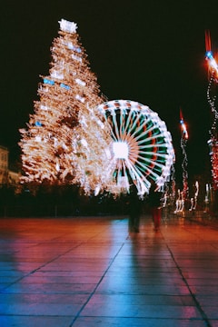 A large, illuminated Christmas tree is adorned with numerous lights, standing next to a brightly lit Ferris wheel. The scene is captured at night, creating a festive and vibrant atmosphere. The pavement reflects the colorful lights from the decorations, and a couple of blurred figures are walking nearby.