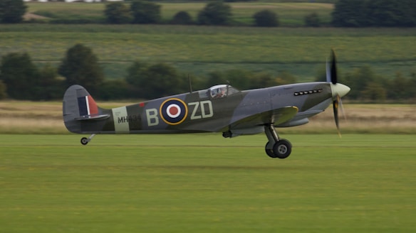 A vintage military aircraft is captured in mid-flight above a grassy landscape, surrounded by fields and distant trees. The aircraft displays military insignia and a propeller in motion, suggesting it is a classic fighter plane.