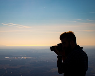 A silhouette of a person holding a camera is captured against a backdrop of a vast landscape and a serene sky. The image features soft lighting from a setting or rising sun, casting a golden hue across the horizon.