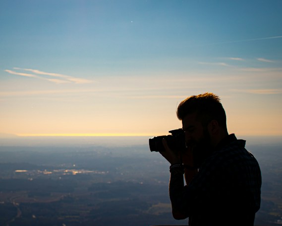 A silhouette of a person holding a camera is captured against a backdrop of a vast landscape and a serene sky. The image features soft lighting from a setting or rising sun, casting a golden hue across the horizon.