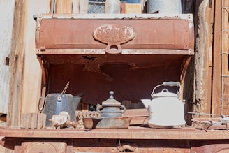 A rusty, old stove with various vintage kitchen items placed on top, including a black kettle, a white teapot, and a metal pot with a lid. The stove's surface is covered with rust and the background features weathered wood and metal textures.