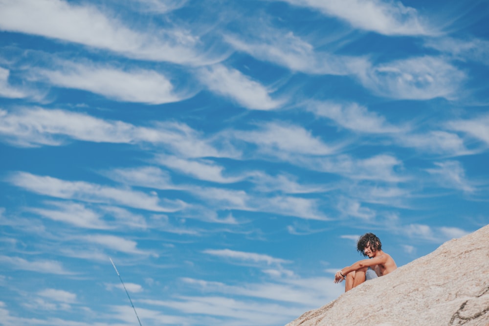 Frau im schwarzen Bikini sitzt tagsüber auf Felsen unter blauem Himmel und weißen Wolken