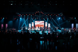 A large stage with people performing under bright stage lights and a central screen displaying colorful visuals. The audience is gathered in the foreground, with blue and red lights illuminating the scene.