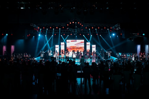 A large stage with people performing under bright stage lights and a central screen displaying colorful visuals. The audience is gathered in the foreground, with blue and red lights illuminating the scene.