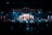 A large stage with people performing under bright stage lights and a central screen displaying colorful visuals. The audience is gathered in the foreground, with blue and red lights illuminating the scene.