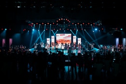 A large stage with people performing under bright stage lights and a central screen displaying colorful visuals. The audience is gathered in the foreground, with blue and red lights illuminating the scene.