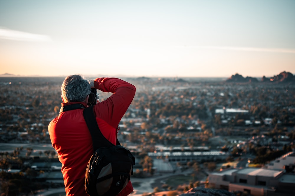 man in red and black jacket sitting on rock looking at city buildings during daytime