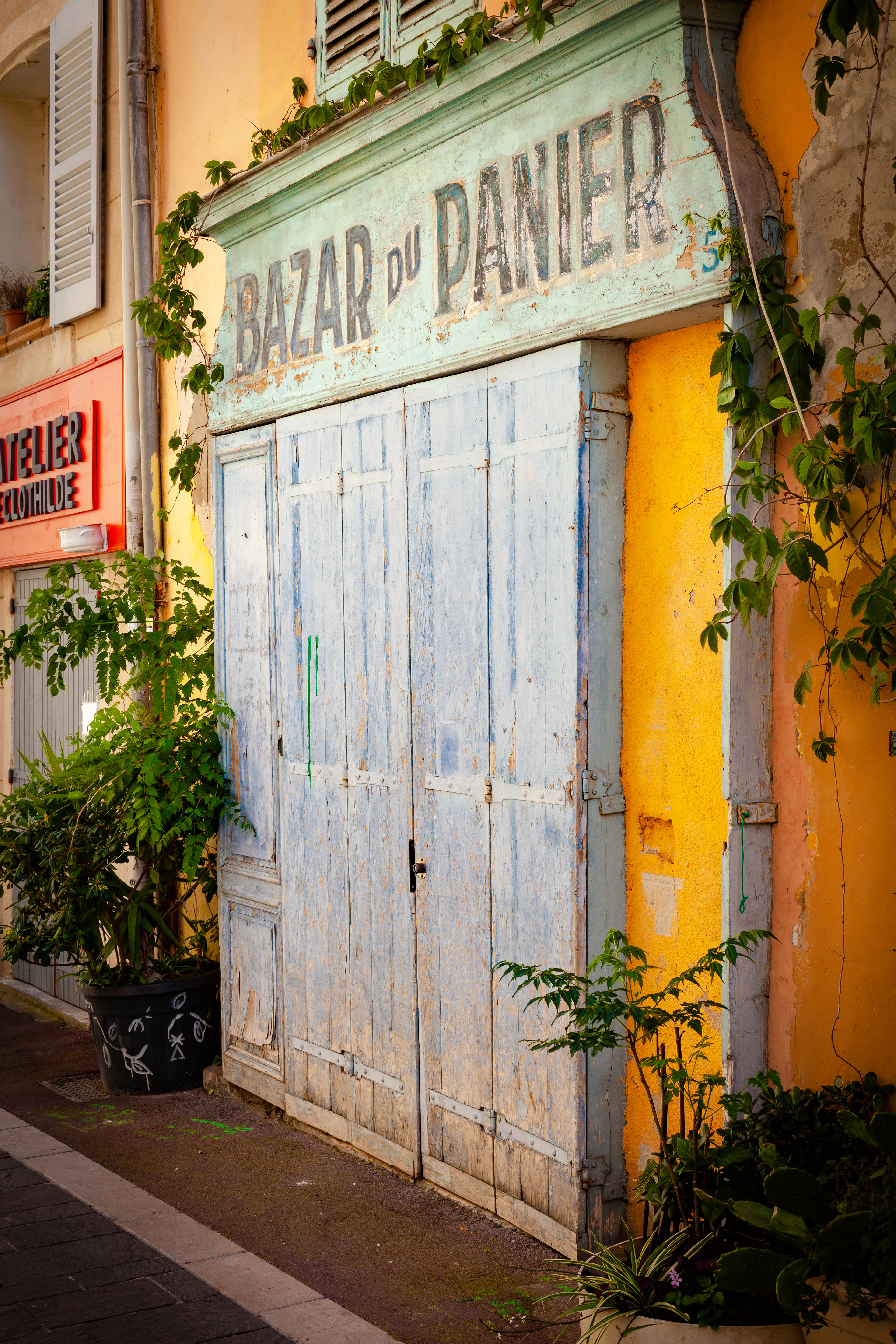 A weathered shopfront in Le Panier, Marseille: pale blue wooden shutters under a mint-green board hand-painted BAZAR DU PANIER, set into a wall of yellow and ochre render with vine leaves spilling down one side.