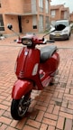 A red scooter is parked in a brick-paved courtyard in front of residential buildings. In the background, there's a silver car with a visible license plate. The area is surrounded by brick walls and large windows.