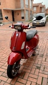 A red scooter is parked in a brick-paved courtyard in front of residential buildings. In the background, there's a silver car with a visible license plate. The area is surrounded by brick walls and large windows.