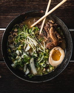 A bowl of ramen with rich broth, topped with crispy pork, half a soft-boiled egg, fresh green onions, sprouts, and sesame seeds is carefully presented, with wooden chopsticks placed on top of the bowl.