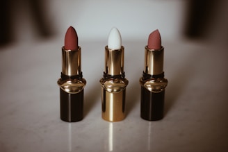 Three lipsticks are arranged in a row on a light surface, each housed in a glossy black and gold tube. The first and third lipsticks have a muted red-pink color, while the middle one is white. The elegant design of the lipstick tubes suggests luxury and sophistication.