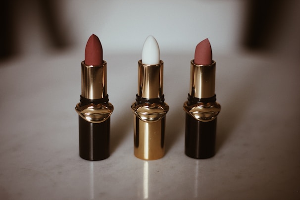 Three lipsticks are arranged in a row on a light surface, each housed in a glossy black and gold tube. The first and third lipsticks have a muted red-pink color, while the middle one is white. The elegant design of the lipstick tubes suggests luxury and sophistication.