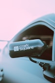 A close-up view of a car's side mirror featuring a carbon fiber housing with the logo 'craft square' printed on it. The mirror is attached to a sleek, modern vehicle, with a soft pink and light blue background, suggesting either an early morning or late afternoon setting.