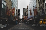 A city street lined with tall buildings and stores, featuring orange banners advertising a comedy festival. Trolley tracks run down the middle of the street, with a tram visible in the distance. The atmosphere appears calm and relatively subdued, with some pedestrians and street furniture visible.