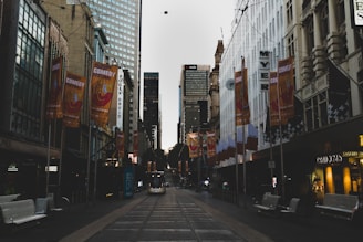 A city street lined with tall buildings and stores, featuring orange banners advertising a comedy festival. Trolley tracks run down the middle of the street, with a tram visible in the distance. The atmosphere appears calm and relatively subdued, with some pedestrians and street furniture visible.