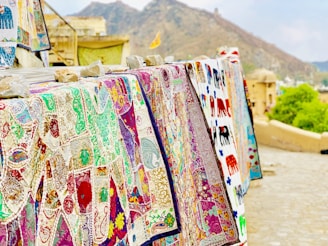 A display of colorful embroidered textiles hanging on a railing. The intricate designs feature various patterns and motifs, including animals and floral elements. The textiles are held down with rocks, and a mountainous landscape is visible in the background.