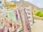 A display of colorful embroidered textiles hanging on a railing. The intricate designs feature various patterns and motifs, including animals and floral elements. The textiles are held down with rocks, and a mountainous landscape is visible in the background.