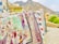 A display of colorful embroidered textiles hanging on a railing. The intricate designs feature various patterns and motifs, including animals and floral elements. The textiles are held down with rocks, and a mountainous landscape is visible in the background.